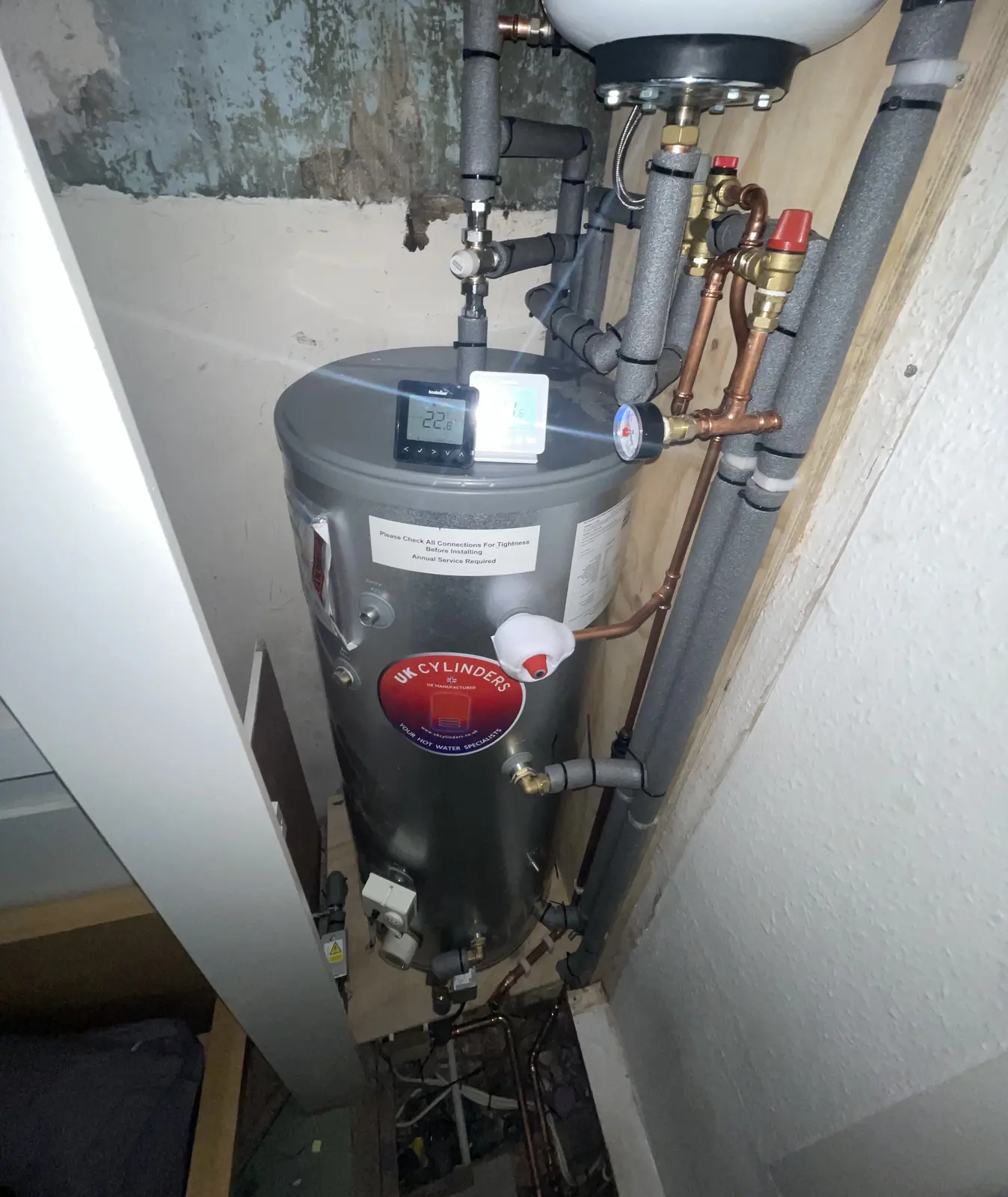 White unvented hot water cylinder in a cupboard, with pipework above it